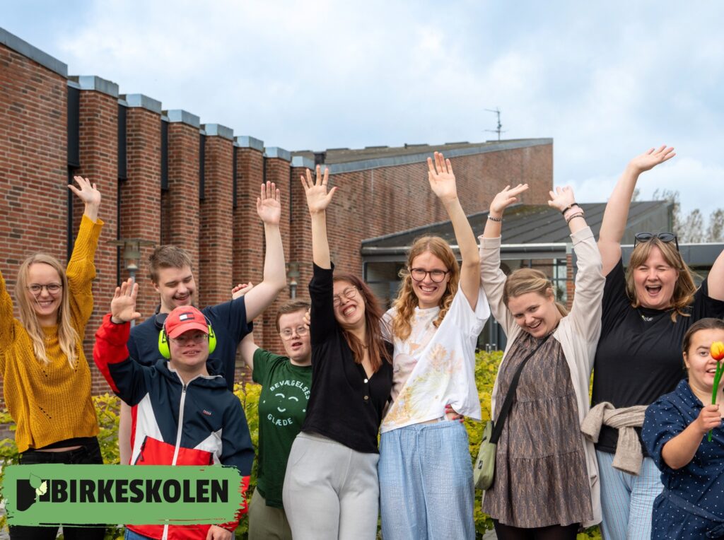 En gruppe mennesker står uden for Birkeskolens lokaler og smiler og rækker hænderne op og fejrer mangfoldigheden. Logoet "BIRKESKOLEN" vises i nederste venstre hjørne.