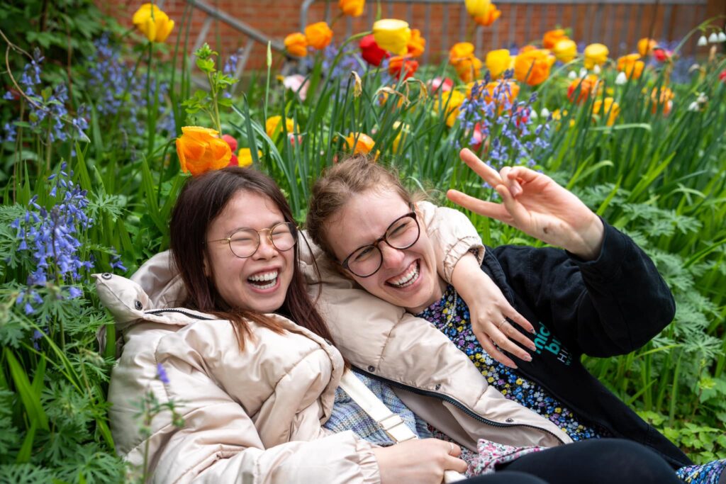 Sophia og Annika sidder i en mark med farverige blomster og smiler og griner, mens Annika laver en fredstegnsbevægelse.