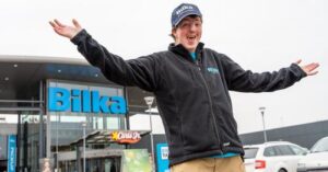 Person i sort jakke og blå kasket stående foran en bilka-butik med bredt spredte arme og et glædeligt udtryk.