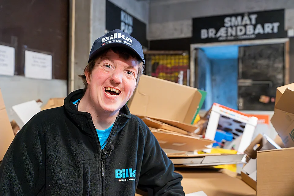 Simon i arbejdstøjet, smilende på et lager fyldt med papkasser.